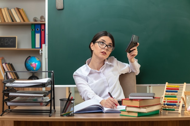 Pensando en buscar joven maestra con gafas sosteniendo calculadora sentado a la mesa con herramientas escolares en el aula