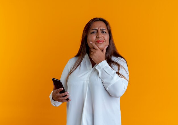 Pensando en casual mujer de mediana edad caucásica sosteniendo el teléfono y poniendo la mano debajo de la barbilla aislada en la pared amarilla