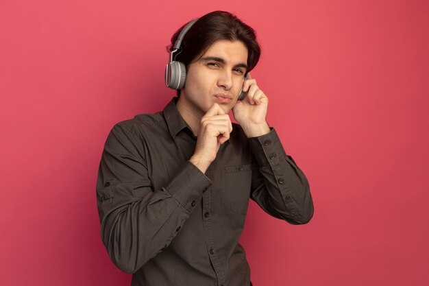 Pensando en el chico guapo joven con camiseta negra con auriculares agarró la barbilla aislada en la pared rosa