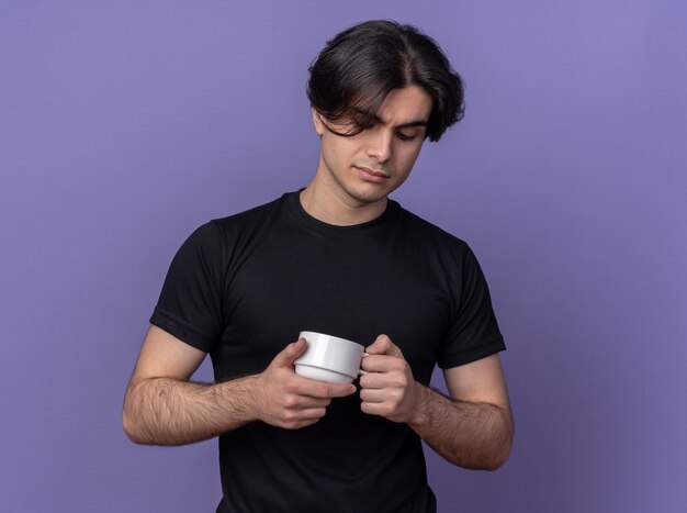 Pensando en chico guapo joven con camiseta negra sosteniendo y mirando una taza de café aislada en la pared púrpura