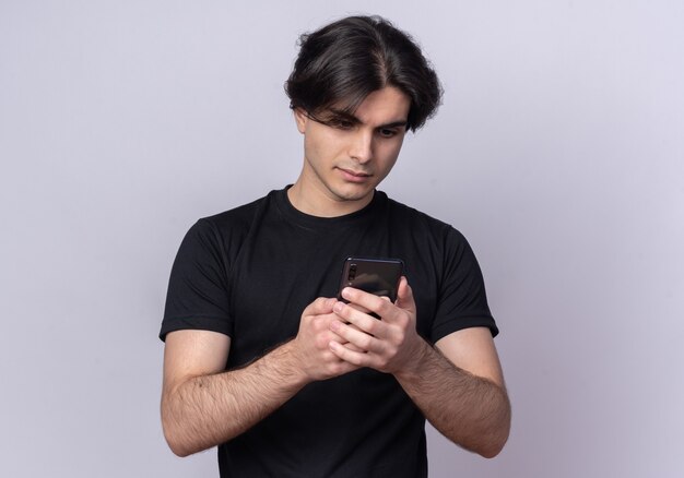 Pensando en chico guapo joven con camiseta negra sosteniendo y mirando el teléfono aislado en la pared blanca