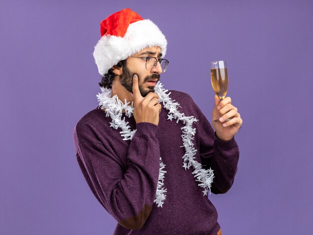 Pensando en chico guapo joven con sombrero de Navidad con guirnalda en el cuello sosteniendo y mirando una copa de champán poniendo el dedo en la mejilla aislado sobre fondo azul.