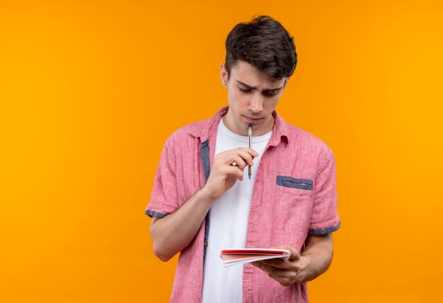 Pensando en chico joven caucásico vistiendo camisa rosa con bolígrafo y cuaderno en pared naranja aislada