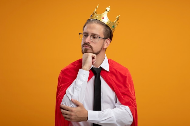 Pensando en el chico joven superhéroe con corbata y corona con gafas poniendo la mano debajo de la barbilla aislada sobre fondo naranja