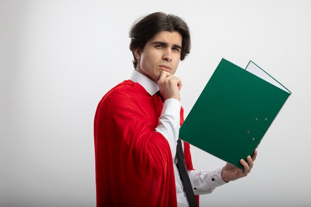 Pensando en chico joven superhéroe con corbata sosteniendo el portapapeles y agarró la barbilla aislado sobre fondo blanco.