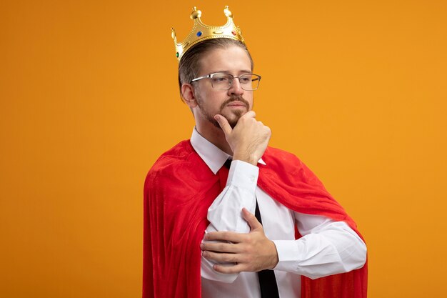 Pensando en el chico joven superhéroe mirando de lado con corbata y corona con gafas agarró la barbilla aislada sobre fondo naranja