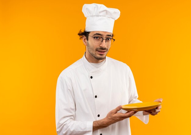 Pensando en el cocinero de sexo masculino joven con uniforme de chef y gafas con placa aislada en la pared amarilla