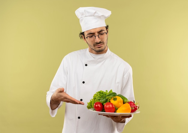Pensando en el cocinero de sexo masculino joven con uniforme de chef y gafas sosteniendo y puntos con verduras de mano en un plato aislado en la pared verde