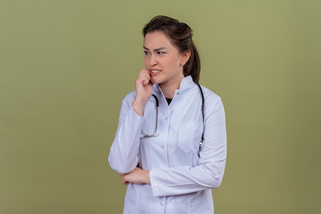 Pensando doctor joven vistiendo bata médica con estetoscopio puso su mano en la mejilla sobre fondo verde