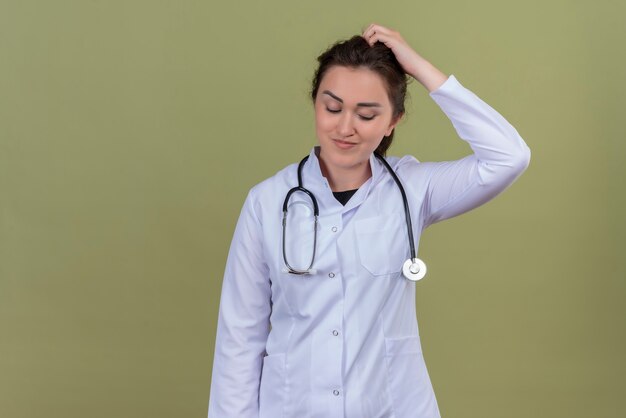 Pensando doctor joven vistiendo bata médica con estetoscopio rasguños sobre fondo verde