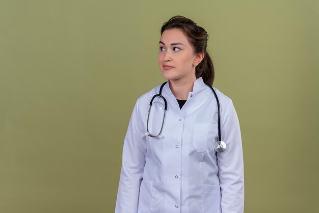 Pensando doctor joven vistiendo bata médica con estetoscopio sobre fondo verde