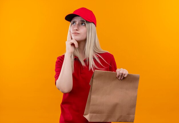 Pensando en la entrega joven vestida con camiseta roja y gorra sosteniendo una caja de papel puso su dedo en la mejilla en la pared naranja aislada