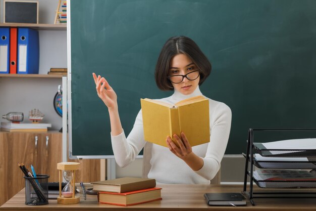 pensando extendiendo las manos joven maestra sosteniendo y leyendo un libro sentado en el escritorio con herramientas escolares en el aula