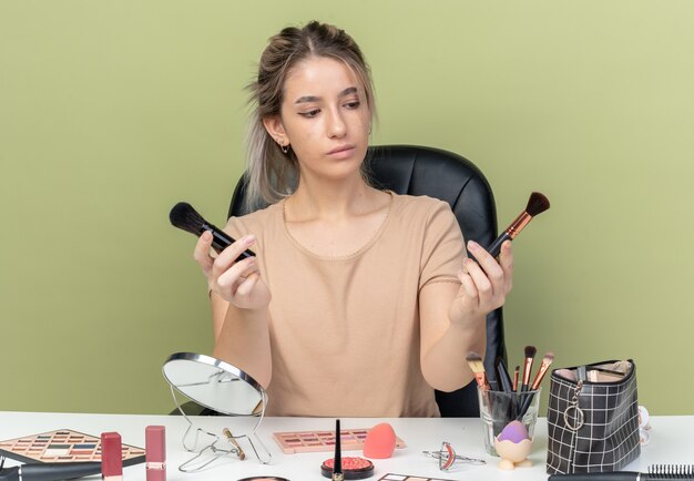 Pensando en la hermosa joven sentada en el escritorio con herramientas de maquillaje sosteniendo y mirando el pincel de maquillaje aislado sobre fondo verde oliva