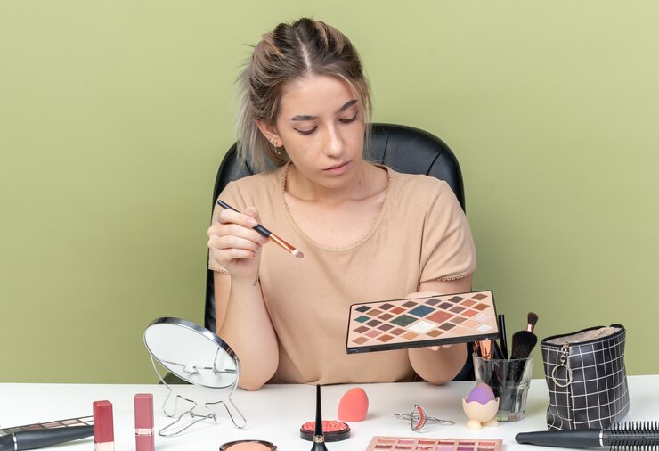 Foto gratuito pensando en la hermosa joven sentada a la mesa con herramientas de maquillaje sosteniendo y mirando el pincel con paleta de sombra de ojos aislado sobre fondo verde oliva