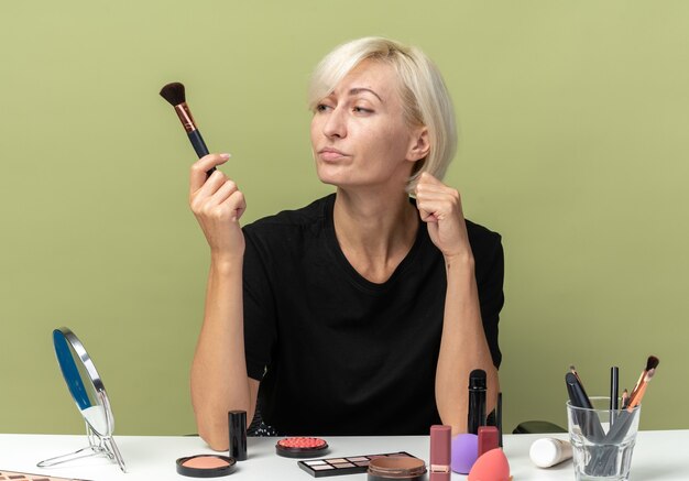 Pensando en la hermosa joven se sienta a la mesa con herramientas de maquillaje sosteniendo y mirando el cepillo de polvo aislado sobre fondo verde oliva