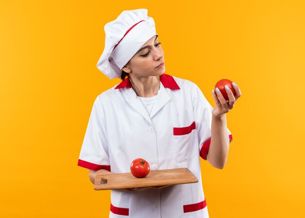 Pensando en la hermosa joven en uniforme de chef sosteniendo y mirando tomates en la tabla de cortar