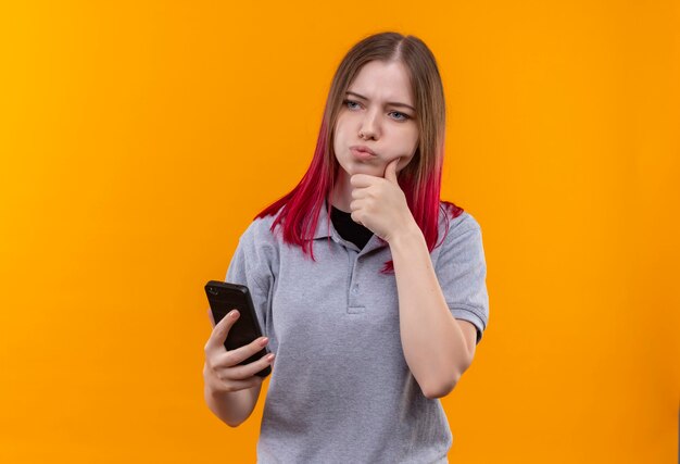 Pensando en la hermosa joven vestida con camiseta gris sosteniendo el teléfono poniendo la mano en la barbilla en la pared amarilla aislada