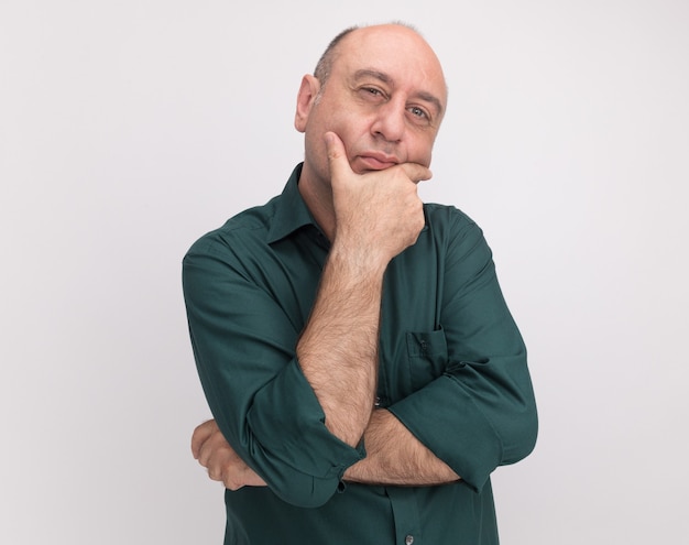 Pensando en el hombre de mediana edad con camiseta verde agarró la barbilla aislada en la pared blanca