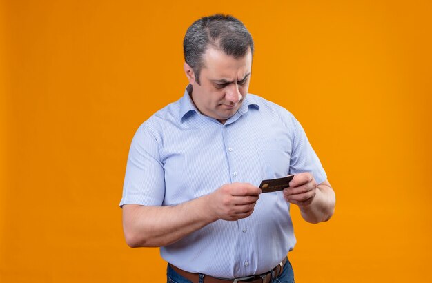 Pensando en el hombre de mediana edad con rayas azules mirando la tarjeta de crédito sobre un fondo naranja