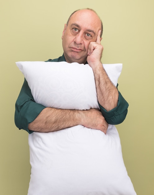 Pensando en el hombre de mediana edad vestido con camiseta verde abrazó la almohada poniendo el dedo en la sien aislado en la pared verde oliva