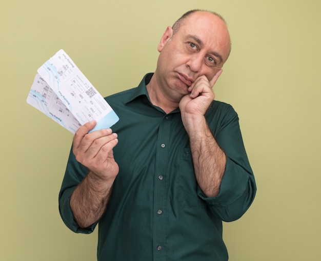 Pensando en inclinar la cabeza hombre de mediana edad vestido con camiseta verde sosteniendo boletos poniendo la mano en la mejilla aislada en la pared verde oliva