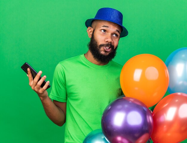 Pensando en el joven afroamericano con sombrero de fiesta sosteniendo globos con teléfono