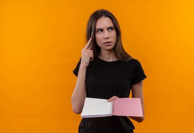 Pensando en la joven caucásica vestida con camiseta negra mirando al lado sosteniendo el cuaderno puso la pluma en la cabeza en la pared naranja aislada