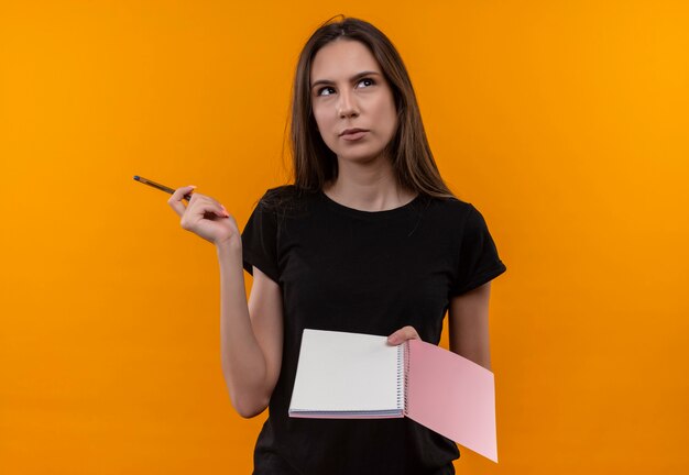 Pensando en la joven caucásica vistiendo camiseta negra con cuaderno y bolígrafo en la pared naranja aislada