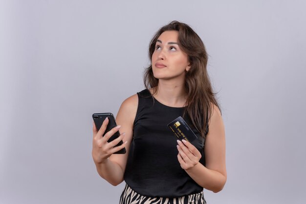 Pensando joven caucásica vistiendo camiseta negra sosteniendo un teléfono y una tarjeta de crédito sobre fondo blanco.