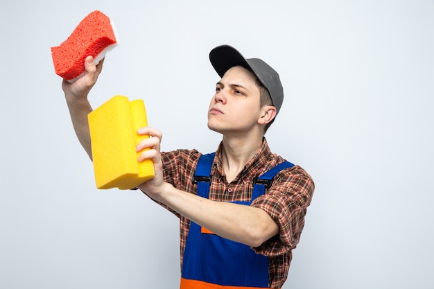 Foto gratuita pensando en el joven chico de limpieza con uniforme y gorra sosteniendo y mirando esponjas