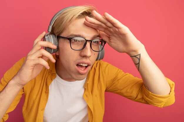 Pensando en el joven chico rubio con camiseta amarilla y gafas con auriculares mirando a distancia con la mano