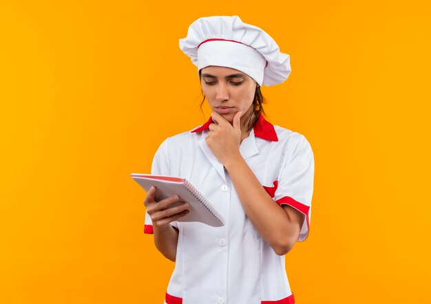 Pensando en la joven cocinera con uniforme de chef sosteniendo y mirando el cuaderno y poniendo la mano debajo de la barbilla con espacio de copia