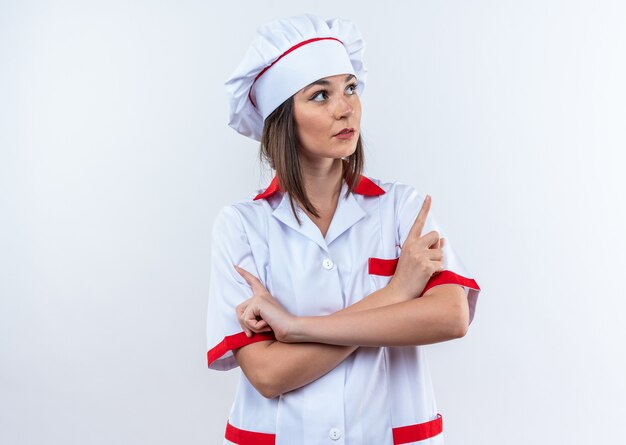 Pensando joven cocinera vistiendo puntos de uniforme de chef en el lado aislado sobre fondo blanco con espacio de copia