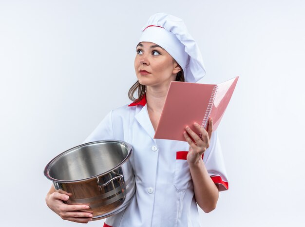 Pensando joven cocinera vistiendo uniforme de chef sosteniendo una cacerola con portátil aislado sobre fondo blanco.