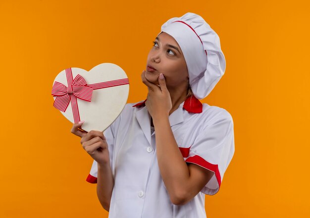 Pensando joven cocinera vistiendo uniforme de chef sosteniendo una caja con forma de corazón poniendo su mano en la barbilla sobre fondo naranja aislado