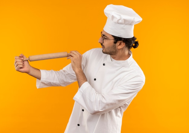 Pensando en el joven cocinero con uniforme de chef y gafas sosteniendo y mirando el rodillo aislado en la pared amarilla