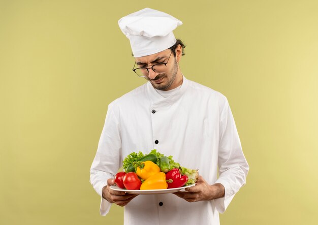 Pensando joven cocinero vistiendo uniforme de chef y gafas sosteniendo y mirando verduras en un plato aislado en la pared verde