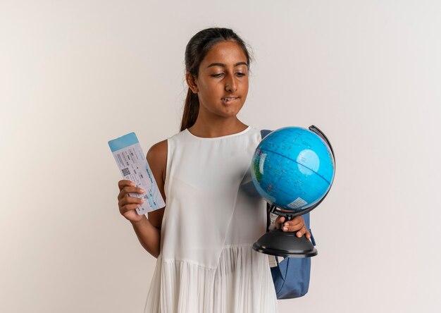 Pensando en la joven colegiala con bolsa trasera sosteniendo el boleto y mirando el globo en su mano aislado en la pared blanca