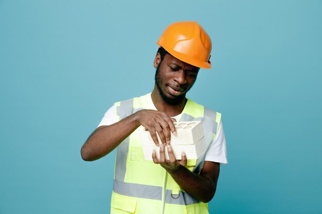 Pensando en un joven constructor afroamericano en uniforme sosteniendo ladrillos aislados de fondo azul