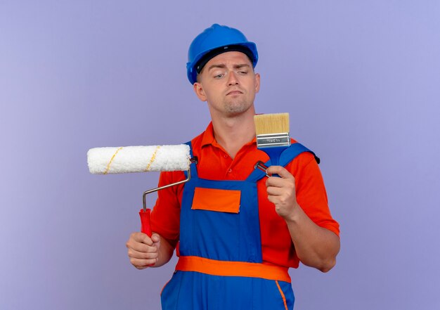 Pensando en el joven constructor con uniforme y casco de seguridad sosteniendo el rodillo de pintura y mirando el pincel en su mano en púrpura