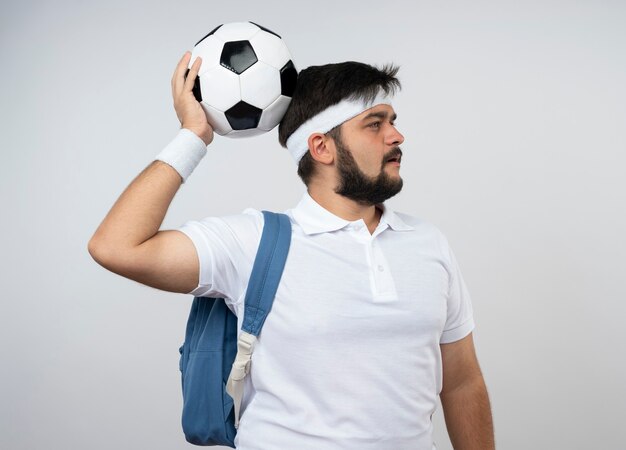 Pensando en el joven deportivo mirando de lado con diadema y muñequera con mochila poniendo la bola en la cabeza aislada en la pared blanca