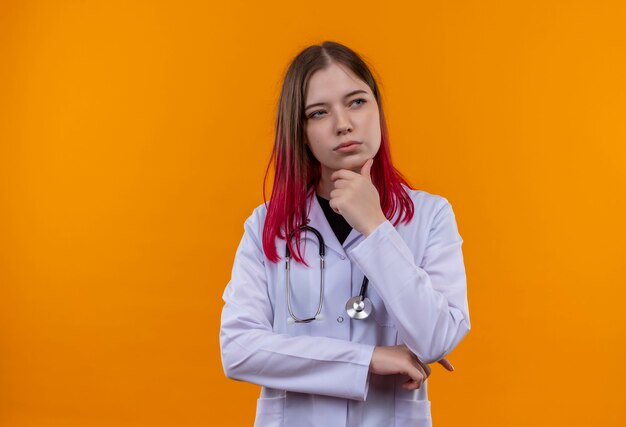 Pensando en la joven doctora vestida con estetoscopio bata médica poniendo la mano en la barbilla en la pared naranja aislada