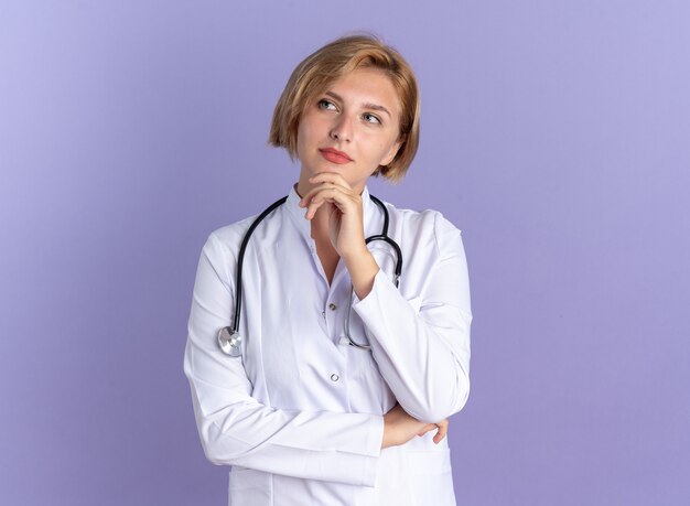 Pensando en la joven doctora vistiendo bata médica con estetoscopio agarró la barbilla aislado sobre fondo azul.