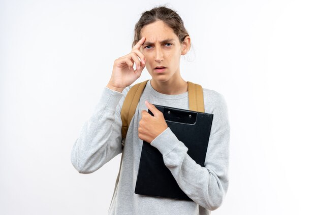 Pensando joven estudiante vistiendo mochila sosteniendo portapapeles poniendo el dedo en la frente aislado en la pared blanca