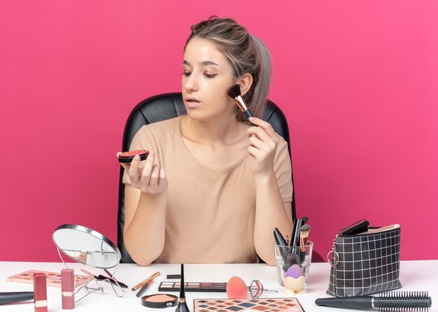 Pensando joven hermosa se sienta a la mesa con herramientas de maquillaje aplicando rubor en polvo aislado sobre fondo rosa