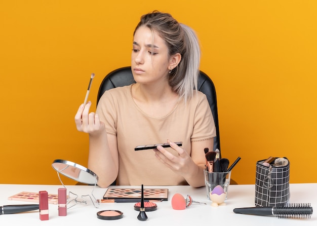 Pensando joven hermosa se sienta a la mesa con herramientas de maquillaje sosteniendo y mirando la paleta de sombra de ojos con pincel de maquillaje aislado sobre fondo naranja