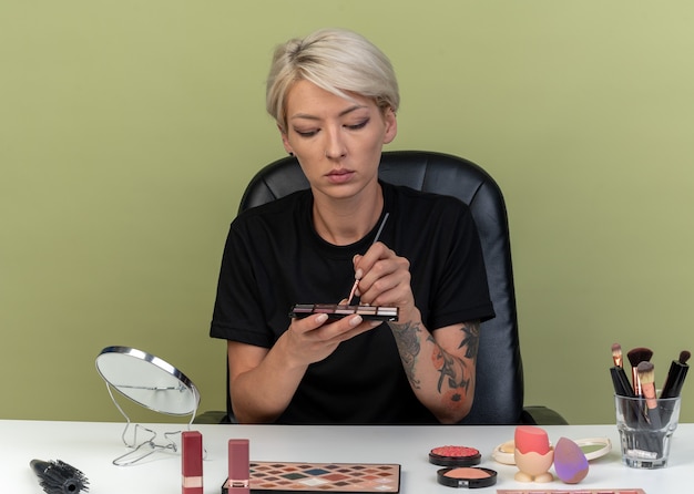 Pensando joven hermosa se sienta a la mesa con herramientas de maquillaje sosteniendo y mirando la paleta de sombras de ojos aislado sobre fondo verde oliva