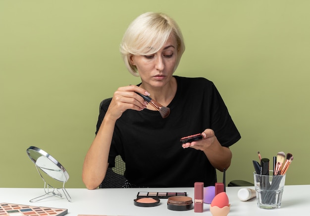 Pensando joven hermosa se sienta a la mesa con herramientas de maquillaje sosteniendo y mirando rubor en polvo aislado sobre fondo verde oliva