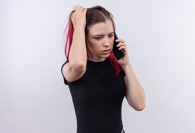 Pensando joven hermosa vestida con camiseta negra habla en el teléfono agarró el pelo en la pared blanca aislada